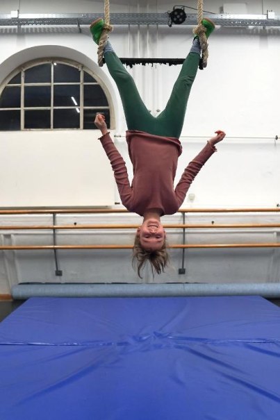 Ein Kind hängt kopfüber an einem Trapez, lächelnd, in einem Turnraum mit blauer Matte und Ballettstange im Hintergrund., © Kunstschule Labyrinth