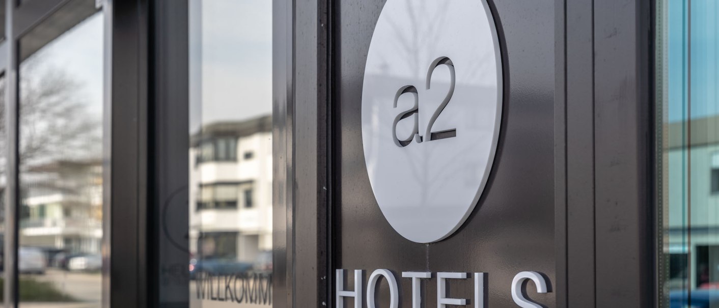 Nahaufnahme des a2 HOTELS Eingangs mit rundem Logo auf der Glasfront. Im Hintergrund sind moderne Gebäude sichtbar., © a2 HOTELS