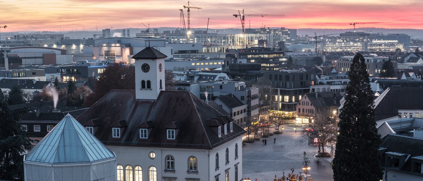 Stadtansicht bei Sonnenuntergang: Ein Kirchturm im Vordergrund, moderne Gebäude und Baukräne im Hintergrund, beleuchtete Fenster und ein farbenfroher Himmel., © Stadt Sindelfingen Stadtansicht bei Sonnenuntergang: Ein Kirchturm im Vordergrund, moderne Gebäude und Baukräne im Hintergrund, beleuchtete Fenster und ein farbenfroher Himmel., © Stadt Sindelfingen