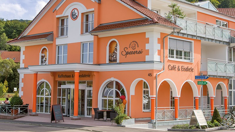 Orangefarbenes Gebäude mit Aufschrift 'Kaffeehaus-Spezerei Sanct Bernhard'. Menschen sitzen draußen an Tischen. Fahrräder stehen vor dem Gebäude., © Kräuterhaus Sanct Bernhard KG Orangefarbenes Gebäude mit Aufschrift 'Kaffeehaus-Spezerei Sanct Bernhard'. Menschen sitzen draußen an Tischen. Fahrräder stehen vor dem Gebäude., © Kräuterhaus Sanct Bernhard KG
