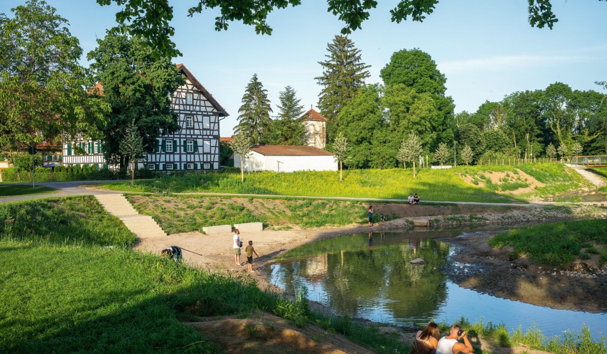 Fachwerkhaus neben einem kleinen Fluss, umgeben von grüner Landschaft. Menschen entspannen am Ufer unter blauem Himmel., © Remstal Tourismus e.V. Fachwerkhaus neben einem kleinen Fluss, umgeben von grüner Landschaft. Menschen entspannen am Ufer unter blauem Himmel., © Remstal Tourismus e.V.