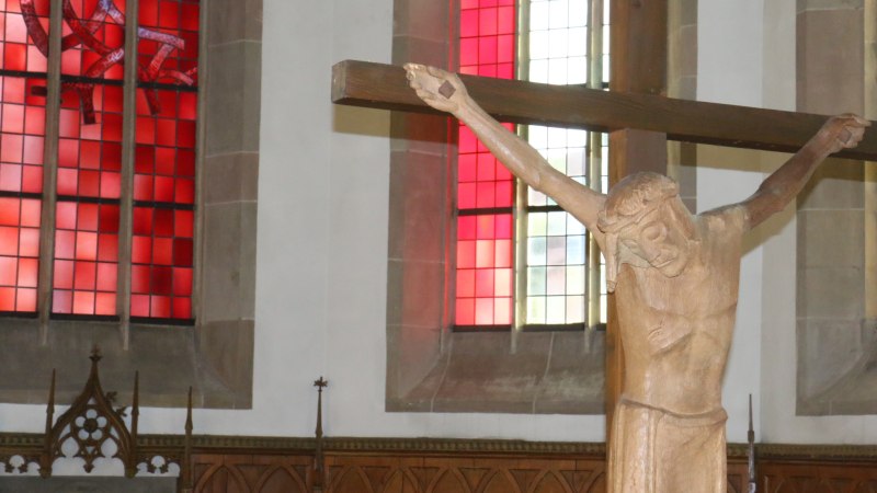 Holzkreuz mit Jesusfigur vor einem roten und bunten Kirchenfenster in der Stadtkirche Gaildorf., &copy; Petra Natzkowski
