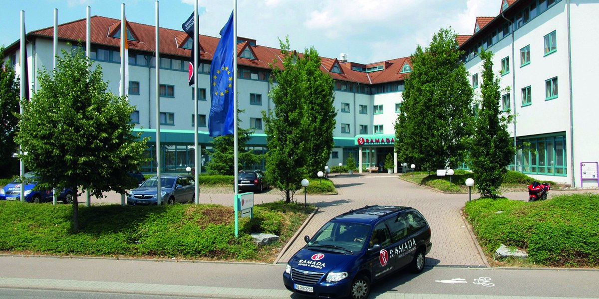 Außenansicht eines Hotels mit Ramada-Logo, umgeben von Bäumen und Flaggen. Ein Ramada-Auto steht im Vordergrund auf der Zufahrt., © © H-Hotels.com