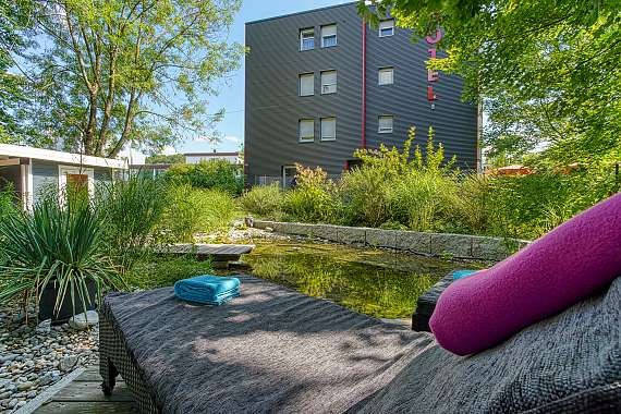 Ein moderner Garten mit Teich, umgeben von Pflanzen. Im Vordergrund ein Liegestuhl mit Handtüchern, im Hintergrund ein mehrstöckiges Gebäude., © Centro Hotels