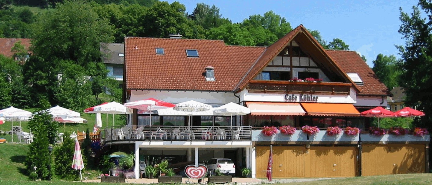 Café Köhler mit rotem Dach, Terrasse mit weißen Stühlen und Sonnenschirmen, umgeben von grüner Landschaft., © Café Köhler