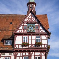 Fachwerk-Rathaus von Bad Urach mit Uhr und Blumenkästen, unter blauem Himmel. Das Gebäude hat ein rotes Dach und eine Turmspitze., © Bad Urach Tourismus