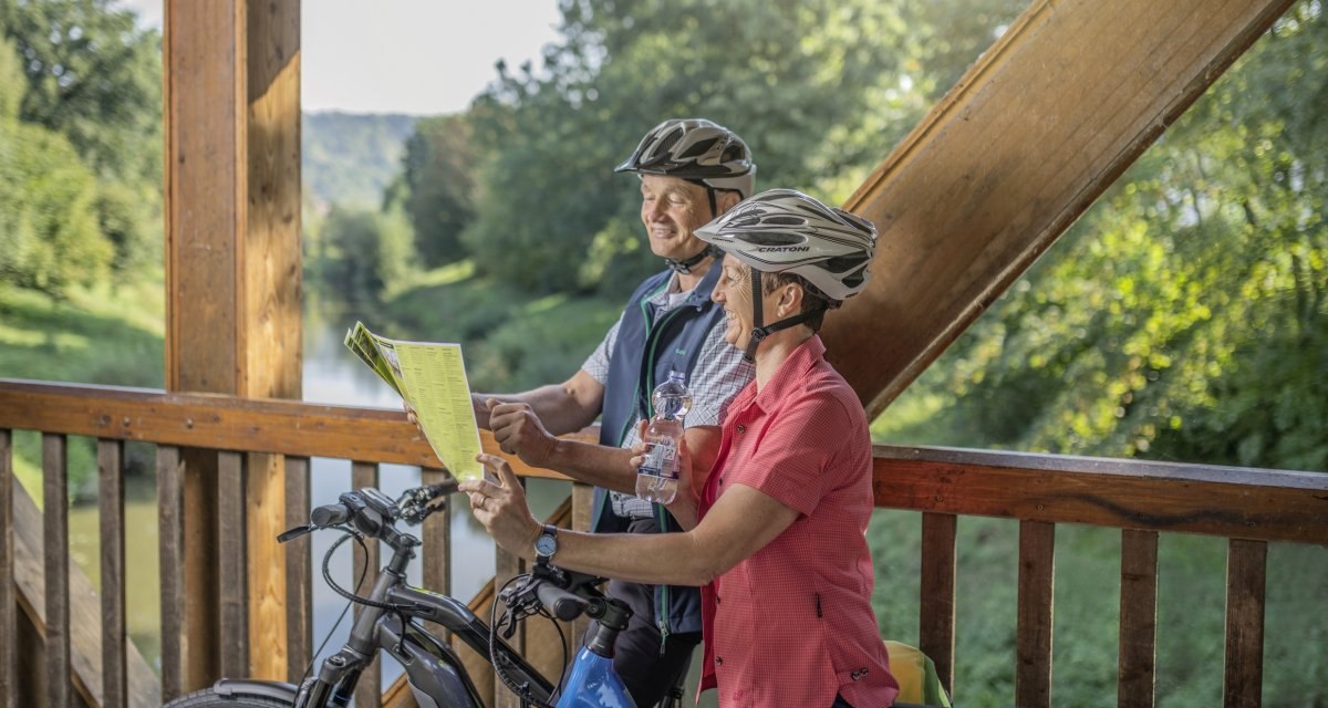 Zwei Radfahrer mit Helmen stehen auf einer Holzbrücke und betrachten eine Karte. Im Hintergrund ist grüne Landschaft zu sehen., © Rems-Murr-Kreis, Jan Bürgermeister Zwei Radfahrer mit Helmen stehen auf einer Holzbrücke und betrachten eine Karte. Im Hintergrund ist grüne Landschaft zu sehen., © Rems-Murr-Kreis, Jan Bürgermeister