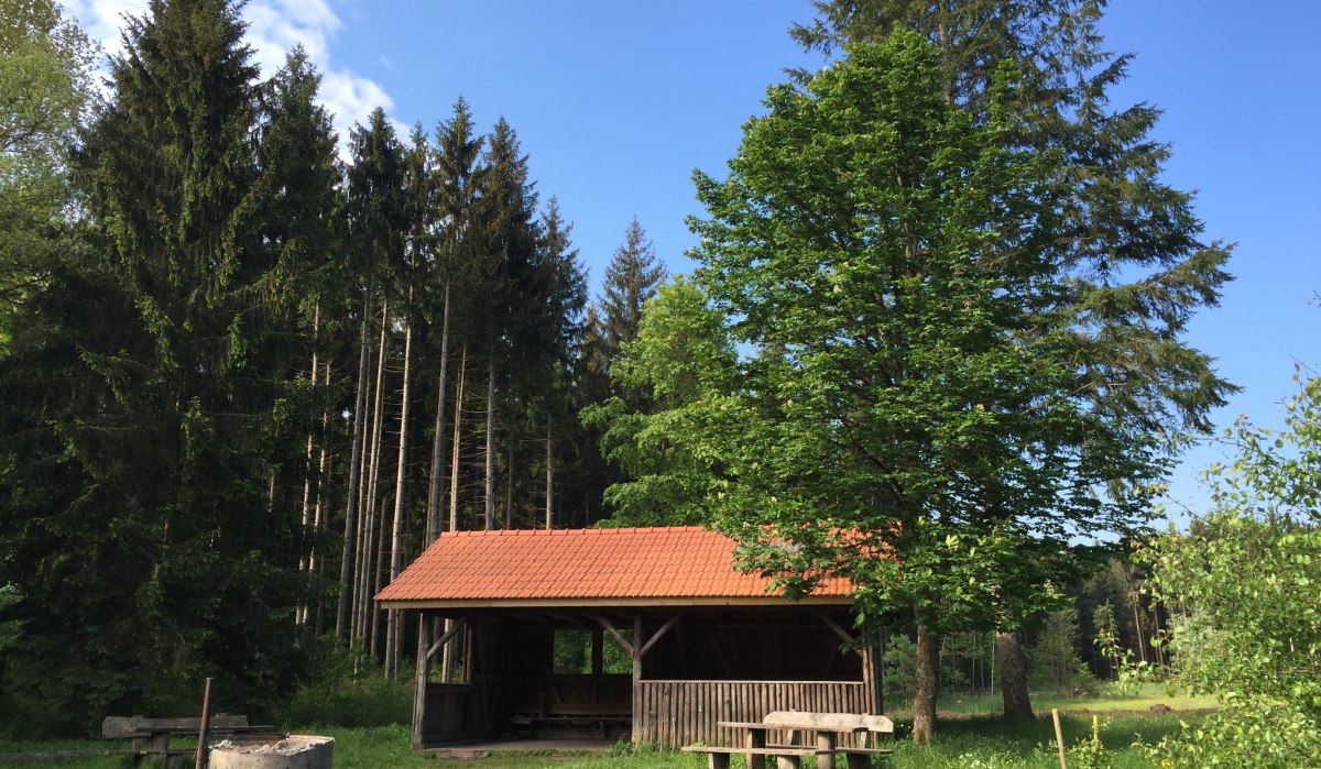 Holzhütte mit rotem Dach in einem Waldgebiet, umgeben von hohen Bäumen und einer Feuerstelle im Vordergrund., © www.pro-cycl.de Holzhütte mit rotem Dach in einem Waldgebiet, umgeben von hohen Bäumen und einer Feuerstelle im Vordergrund., © www.pro-cycl.de