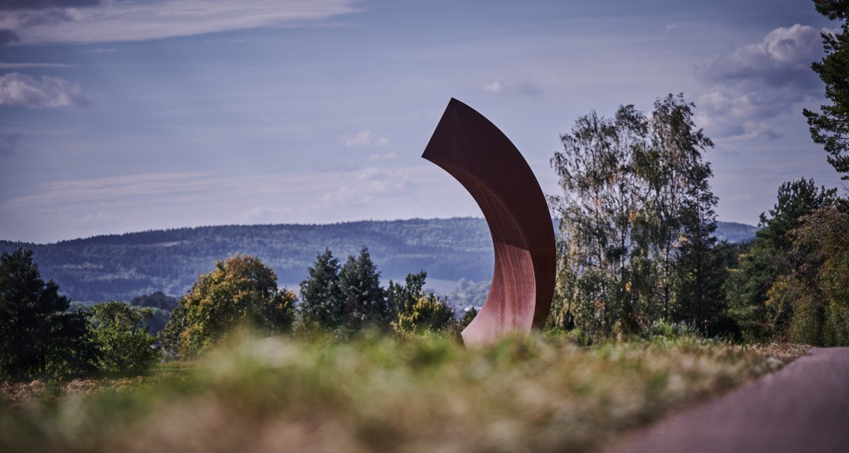 Gro&szlig;e, gebogene Skulptur in einer Landschaft mit B&auml;umen und H&uuml;geln im Hintergrund unter blauem Himmel., &copy; Natur.Nah. Sch&ouml;nbuch & Heckeng&auml;u