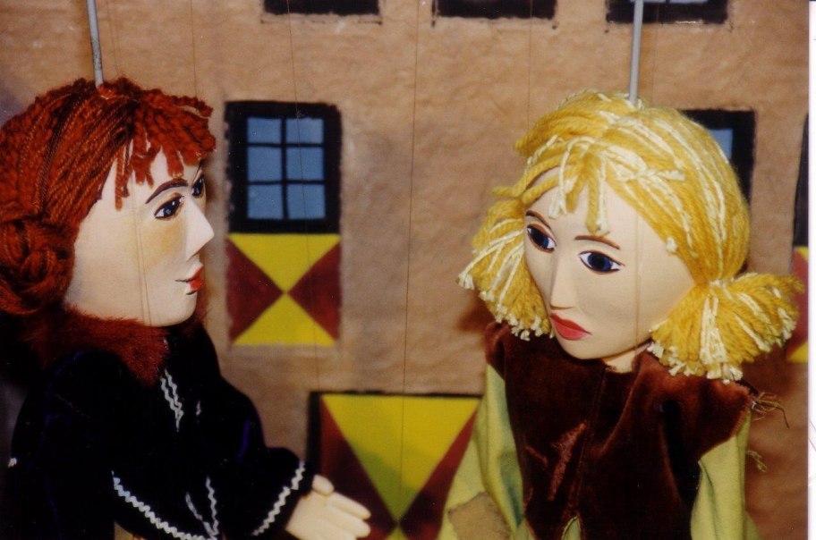 Zwei Marionetten mit roten und blonden Haaren stehen vor einem bemalten Hintergrund mit Fenstern und geometrischen Mustern., © Stadtbücherei Bad Urach