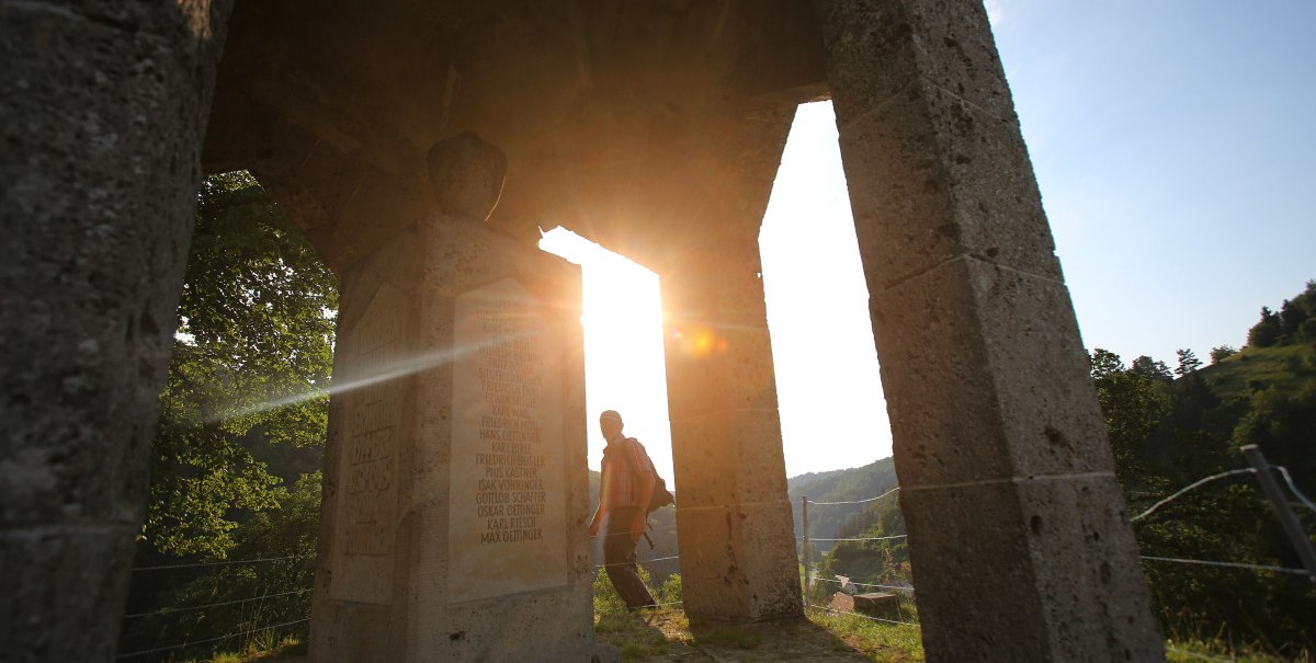 Ein Ehrenmal auf einem Hügel, beleuchtet von der untergehenden Sonne. Eine Person steht im Hintergrund, umgeben von Natur., © Bad Urach Tourismus