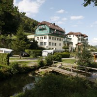 Ein Resort mit Wohnwagen, Gebäuden und einer Holzbrücke in einer grünen, bewaldeten Landschaft unter blauem Himmel., © Family Resort Kleinenzhof Blick in den Nord Schwarzwald Ein Resort mit Wohnwagen, Gebäuden und einer Holzbrücke in einer grünen, bewaldeten Landschaft unter blauem Himmel., © Family Resort Kleinenzhof Blick in den Nord Schwarzwald