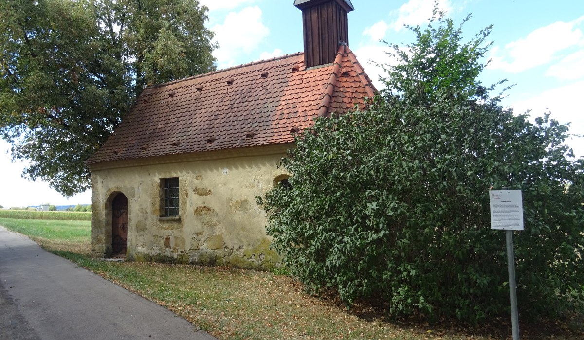 Kleine Kapelle mit rotem Ziegeldach und kleinem Turm, umgeben von Bäumen und Sträuchern. Ein Weg führt an der Kapelle vorbei., © Foto: Cornelia Steinbach Kleine Kapelle mit rotem Ziegeldach und kleinem Turm, umgeben von Bäumen und Sträuchern. Ein Weg führt an der Kapelle vorbei., © Foto: Cornelia Steinbach