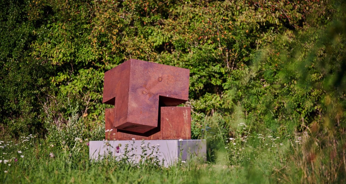 Rostige Metallskulptur auf einem Sockel, umgeben von üppiger Vegetation und Wildblumen. Die Skulptur besteht aus geometrischen Formen., © Natur.Nah. Schönbuch & Heckengäu Rostige Metallskulptur auf einem Sockel, umgeben von üppiger Vegetation und Wildblumen. Die Skulptur besteht aus geometrischen Formen., © Natur.Nah. Schönbuch & Heckengäu