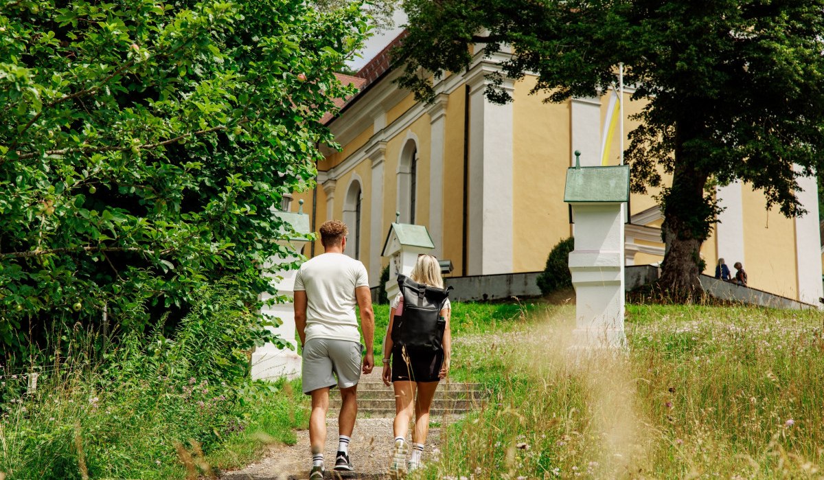 Zwei Personen gehen einen Weg entlang, der zu einer gelben Kirche führt, umgeben von Bäumen und Gras., © Landkreis Göppingen
