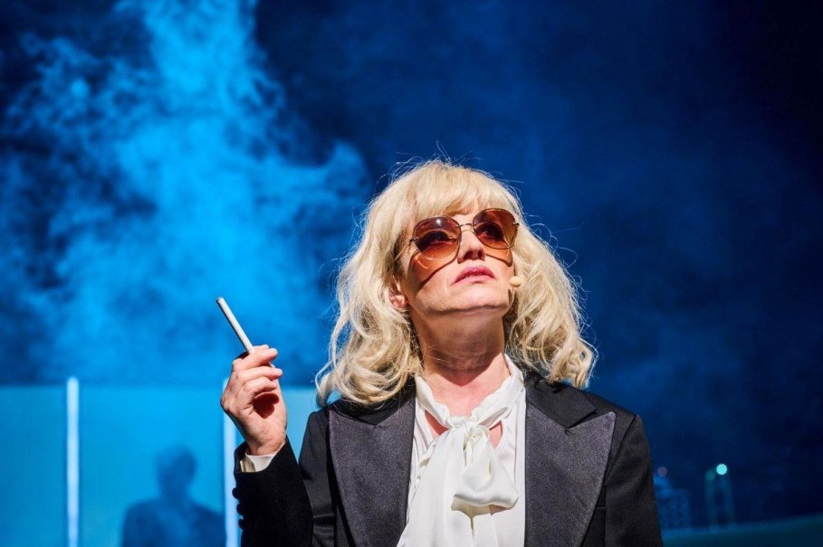 Eine Person mit blonder Per&uuml;cke und Sonnenbrille steht auf einer B&uuml;hne, h&auml;lt eine Zigarette und tr&auml;gt ein Mikrofon. Der Hintergrund ist blau beleuchtet., &copy; W&uuml;rttembergische Landesb&uuml;hne Esslingen