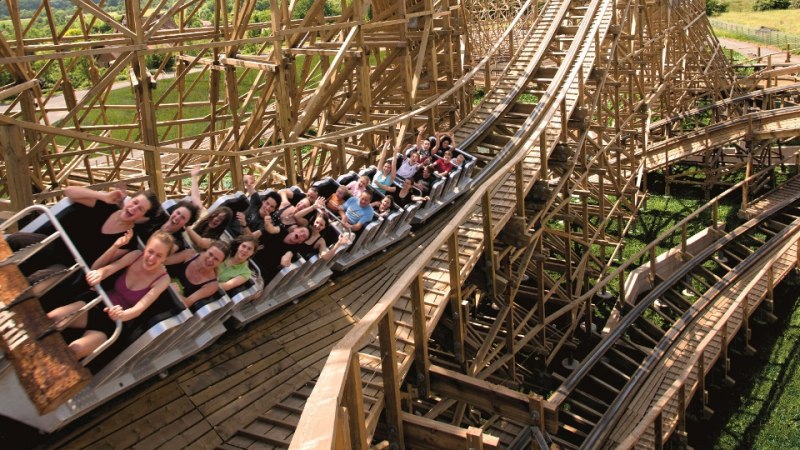 Eine Gruppe von Menschen genie&szlig;t eine Fahrt auf einer Holzachterbahn, w&auml;hrend sie die Arme in die Luft strecken. Die Bahn schl&auml;ngelt sich durch gr&uuml;ne Landschaft., &copy; Erlebnispark Tripsdrill