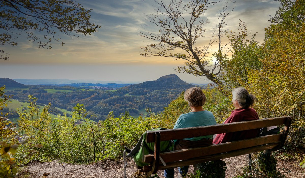 Zwei Personen sitzen auf einer Bank am Aussichtspunkt Luginsland und genießen den Blick auf die hügelige Landschaft im Sonnenuntergang., © Foto: Mario Klaiber