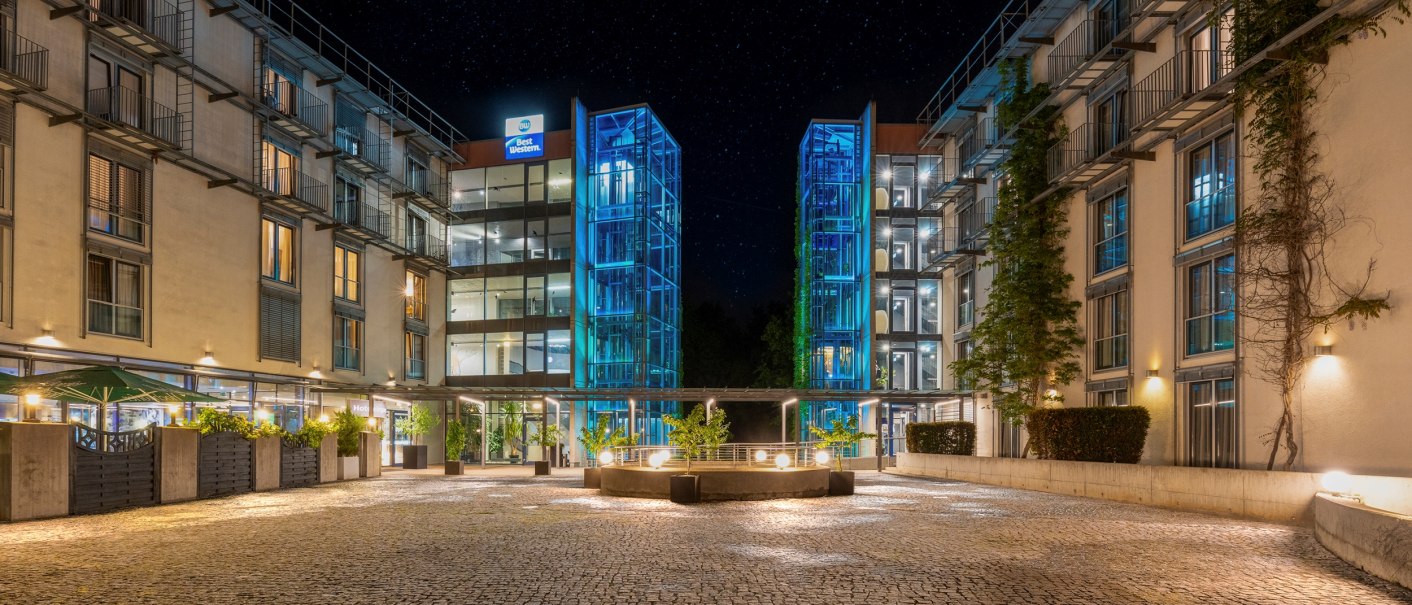 Nachtaufnahme eines modernen Hotels mit beleuchteten Glasaufzügen und einem gepflasterten Innenhof. Sterne sind am Himmel sichtbar., © Plaza Residence Stuttgart-Ditzingen Nachtaufnahme eines modernen Hotels mit beleuchteten Glasaufzügen und einem gepflasterten Innenhof. Sterne sind am Himmel sichtbar., © Plaza Residence Stuttgart-Ditzingen