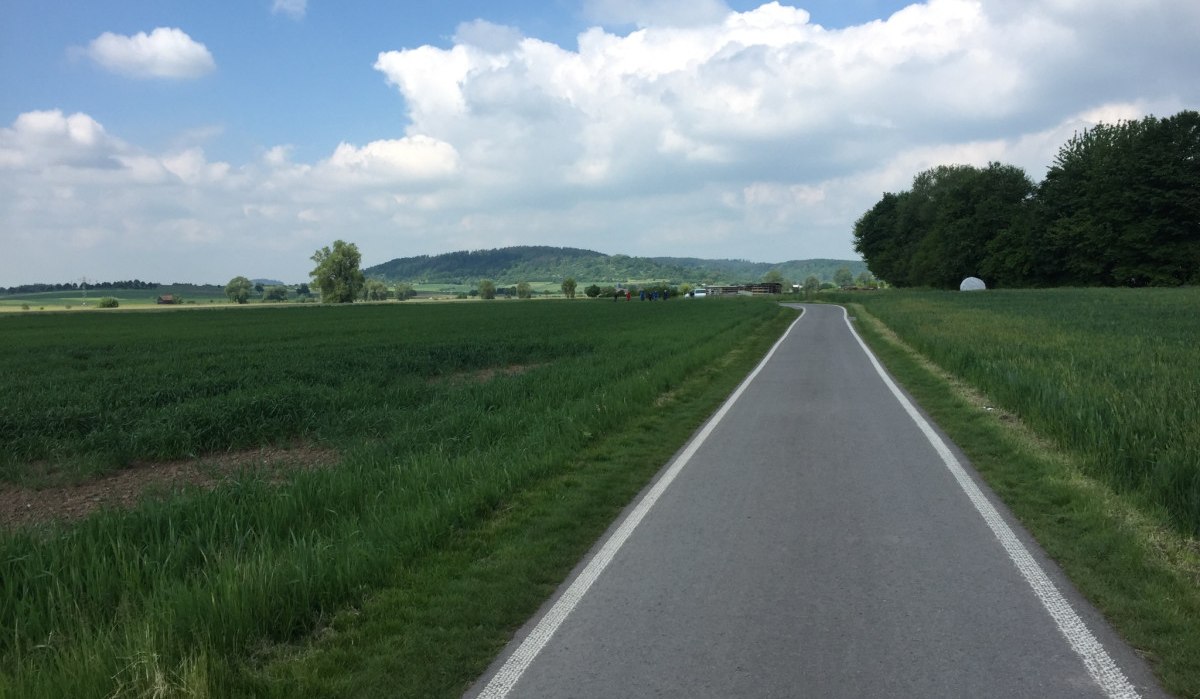 Ein asphaltierter Radweg schlängelt sich durch grüne Felder und Bäume, unter einem blauen Himmel mit weißen Wolken., © www.pro-cycl.de Ein asphaltierter Radweg schlängelt sich durch grüne Felder und Bäume, unter einem blauen Himmel mit weißen Wolken., © www.pro-cycl.de