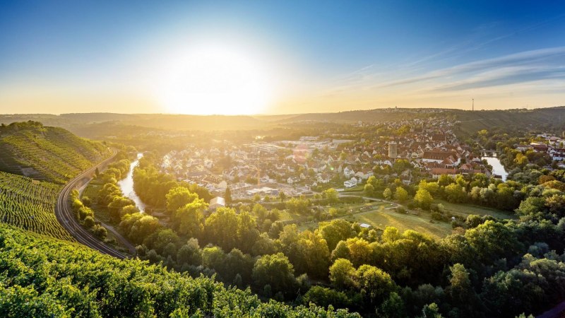 Das Bild zeigt den Blick von der Weinkanzel in Besigheim im Herbst. Das Motiv wird dominiert von Wein, der an den Steillagen angebaut wird. Im Tal ist der an dieser Stelle U-förmig verlaufende Neckar zu sehen. Im Hintergrund erkennt man die braunen Dächer der Stadt. Dieses Motiv nimmt die unteren 2/3 des Bilds ein. Darüber erstreckt sich blauer Himmel mit der tief stehenden Sonne., © Stuttgart-Marketing GmbH, Sarah Schmid