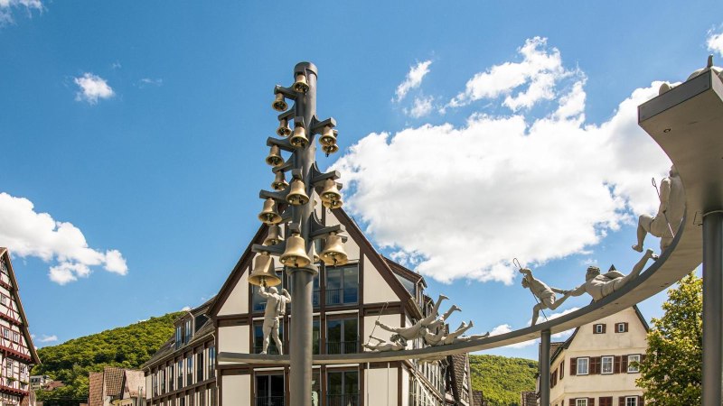 Skulptur mit Glocken und Figuren auf dem Marktplatz von Bad Urach, umgeben von Fachwerkhäusern und blauem Himmel., © Stuttgart-Marketing GmbH, Sarah Schmid