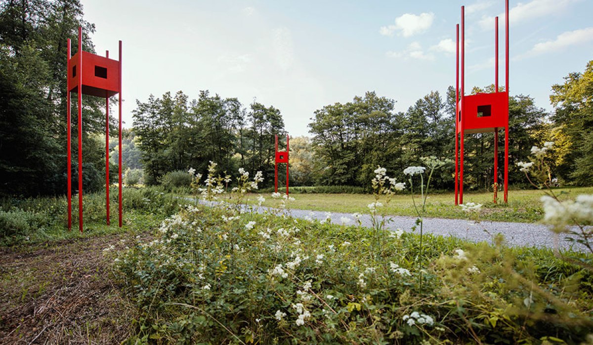 Rote Skulpturen mit quadratischen Elementen stehen auf hohen Stangen in einer Waldlandschaft. Weiße Blumen blühen im Vordergrund., © Natur.Nah. Schönbuch & Heckengäu Rote Skulpturen mit quadratischen Elementen stehen auf hohen Stangen in einer Waldlandschaft. Weiße Blumen blühen im Vordergrund., © Natur.Nah. Schönbuch & Heckengäu