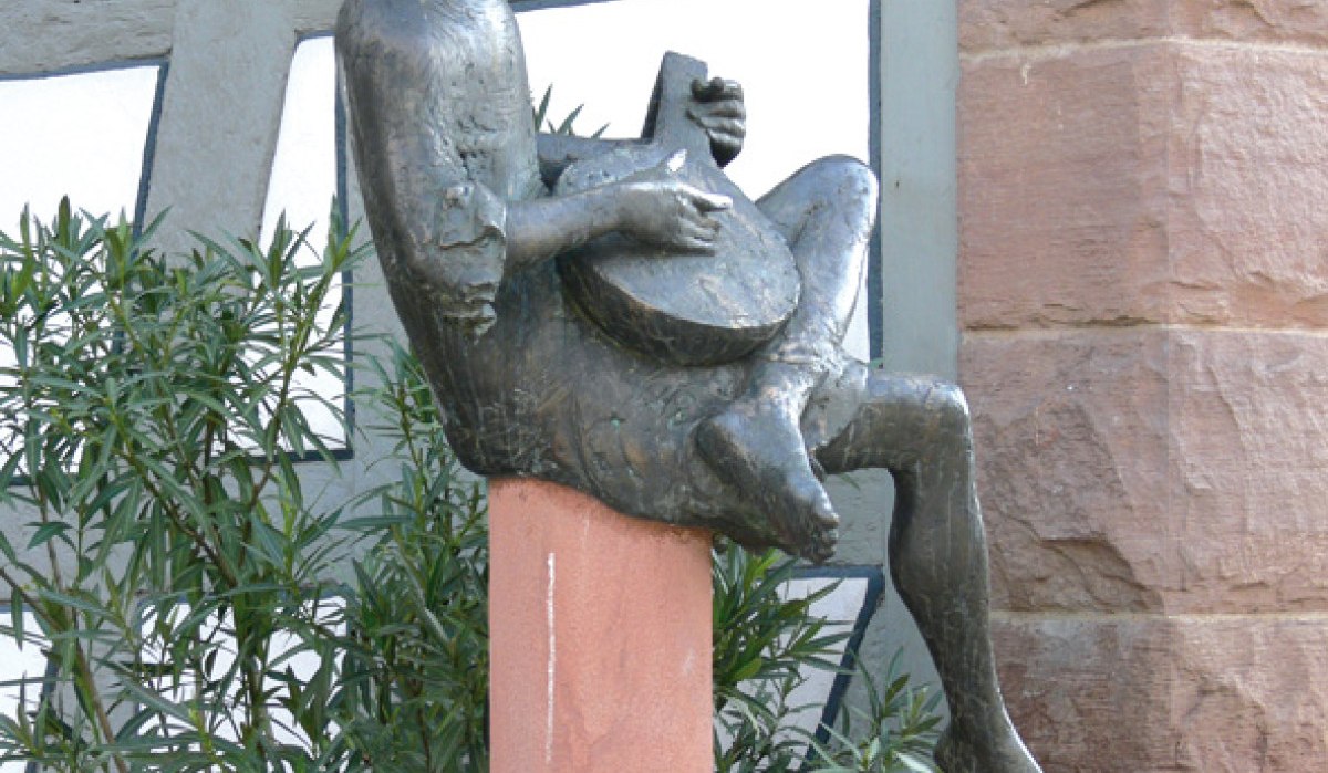 Bronzestatue eines Minnesängers mit Laute, sitzend auf einem Sockel vor einem Fachwerkgebäude. Im Hintergrund sind Pflanzen und eine Steinmauer zu sehen., © Land der 1000 Hügel - Kraichgau-Stromberg Bronzestatue eines Minnesängers mit Laute, sitzend auf einem Sockel vor einem Fachwerkgebäude. Im Hintergrund sind Pflanzen und eine Steinmauer zu sehen., © Land der 1000 Hügel - Kraichgau-Stromberg