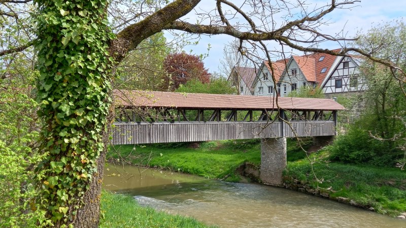 Fluss mit überdachter Holzbrücke, am Ufer Büsche und Bäume, auf der rechten Bildseite sind die weißen Giebelseiten und rotbraunen Dachflächen von vier Häusern hinter der Holzbrücke zu erkennen, © Stadt Gaildorf
