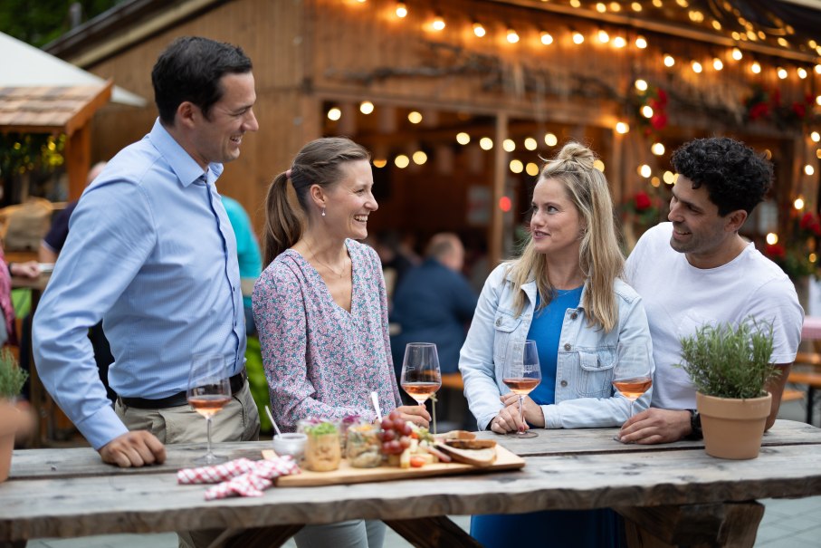 Vier Personen lachen und unterhalten sich an einem Holztisch mit Wein und Snacks auf einem Weinfest. Im Hintergrund sind Lichter und eine Holzhütte zu sehen., © Stadt Nürtingen, B. Knoblauch