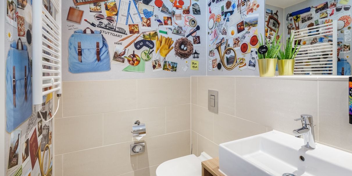 Modernes Badezimmer mit Collage-Wand, weißen Fliesen, Waschbecken und Toilette. Pflanzen in goldenen Töpfen als Dekoration., © Novum Hospitality Modernes Badezimmer mit Collage-Wand, weißen Fliesen, Waschbecken und Toilette. Pflanzen in goldenen Töpfen als Dekoration., © Novum Hospitality