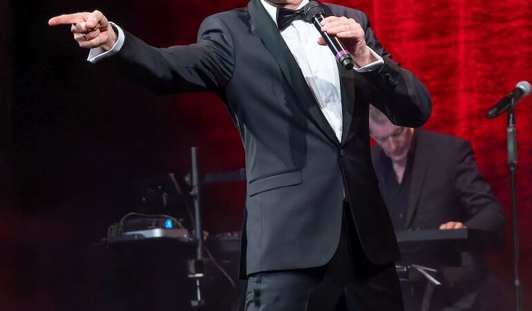 Ein Mann im Anzug und mit Sonnenbrille performt auf einer B&uuml;hne vor einem roten Vorhang. Er h&auml;lt ein Mikrofon und zeigt mit dem Finger nach vorne., &copy; Stadthalle Leonberg