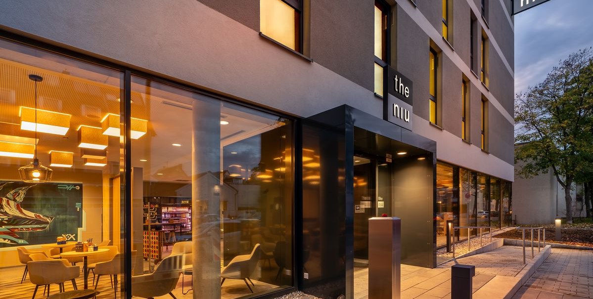 Moderner Hoteleingang mit Glasfassade, beleuchtetem Innenbereich und dem Schriftzug 'the niu'. Außenbereich mit Gehweg und Pflanzen., © Novum Hospitality Moderner Hoteleingang mit Glasfassade, beleuchtetem Innenbereich und dem Schriftzug 'the niu'. Außenbereich mit Gehweg und Pflanzen., © Novum Hospitality