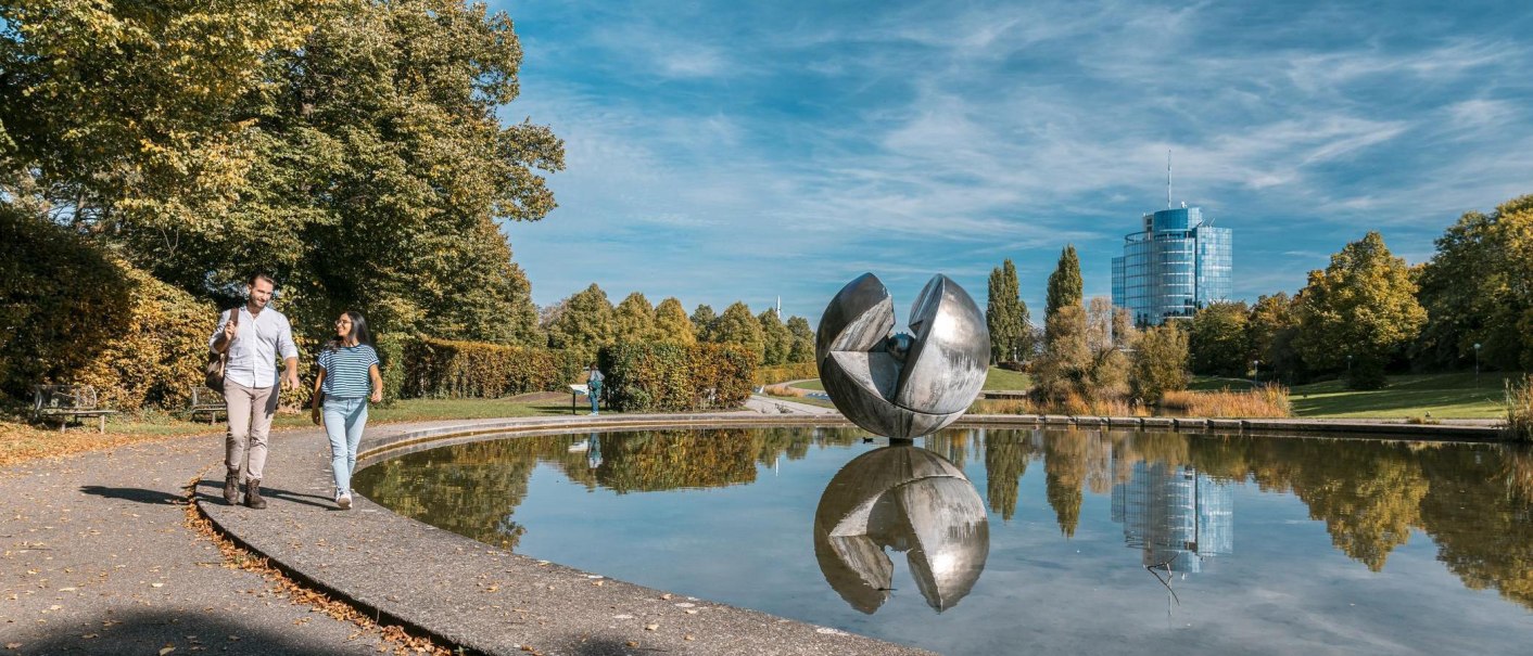 Ein Paar spaziert im Wartbergpark entlang eines Teiches mit einer großen Skulptur und einem modernen Gebäude im Hintergrund., © Stuttgart-Marketing GmbH, Sarah Schmid