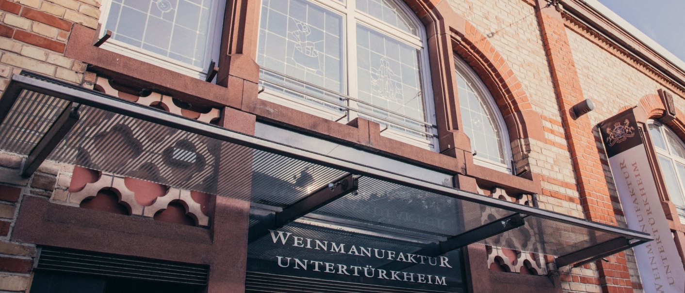 Fassade der Weinmanufaktur Untertürkheim mit Glasvordach, Schriftzug und dekorativen Fenstern., © Weinmanufaktur Stuttgart Fassade der Weinmanufaktur Untertürkheim mit Glasvordach, Schriftzug und dekorativen Fenstern., © Weinmanufaktur Stuttgart