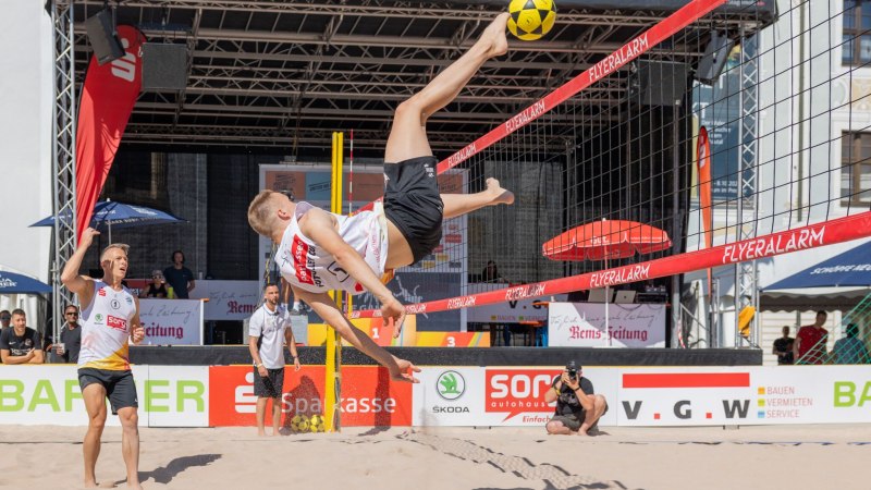 Ein Beachvolleyballspieler führt einen spektakulären Fallrückzieher aus, während sein Teamkollege zuschaut. Im Hintergrund sind Zuschauer und Werbebanner sichtbar., © Für die Bildrechte: Amt für Medien und Kommunikation Schwäbisch Gmünd Ein Beachvolleyballspieler führt einen spektakulären Fallrückzieher aus, während sein Teamkollege zuschaut. Im Hintergrund sind Zuschauer und Werbebanner sichtbar., © Für die Bildrechte: Amt für Medien und Kommunikation Schwäbisch Gmünd