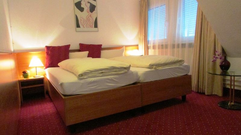 Hotelzimmer mit Doppelbett, rotem Teppich, Nachttischlampe und Bild an der Wand. Fenster mit Vorh&auml;ngen, Tisch mit Vase., &copy; TOMAS
