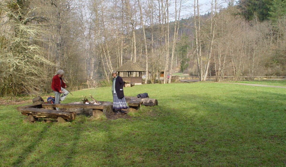Zwei Personen stehen an einer Feuerstelle auf einer Wiese, umgeben von kahlen Bäumen. Im Hintergrund ist eine Holzhütte zu sehen., © Naturpark Schönbuch Zwei Personen stehen an einer Feuerstelle auf einer Wiese, umgeben von kahlen Bäumen. Im Hintergrund ist eine Holzhütte zu sehen., © Naturpark Schönbuch