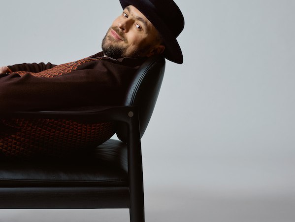 Eine Person mit Hut lehnt entspannt auf einem schwarzen Stuhl, seitlich liegend, vor einem grauen Hintergrund., © Scala Kultur Live gGmbH