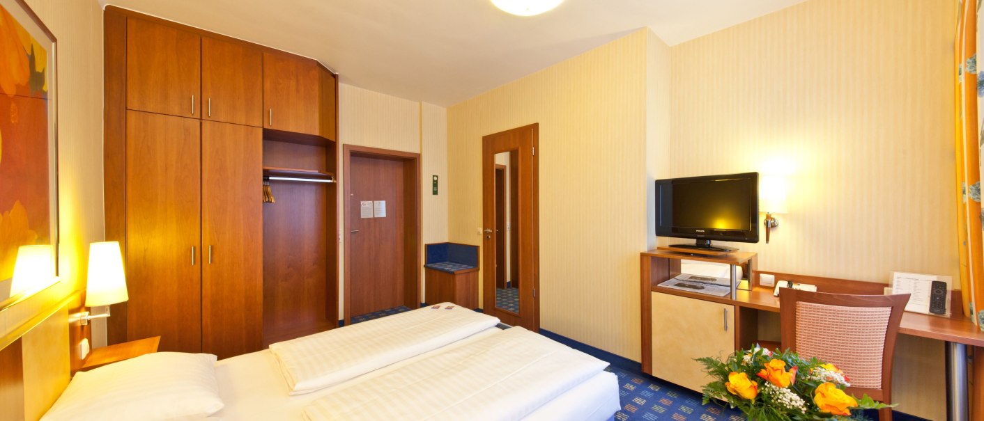 Ein gemütliches Hotelzimmer mit Doppelbett, Schreibtisch, Fernseher und einem Blumenstrauß auf einem Tisch., © Novum Boulevard Stuttgart City