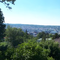 Panoramablick auf eine Stadt mit gr&uuml;nen B&auml;umen im Vordergrund und H&uuml;geln im Hintergrund unter klarem Himmel., &copy; SMG