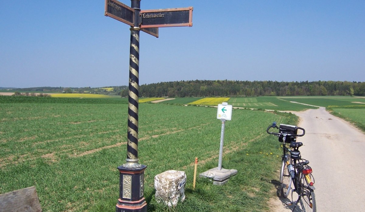 Ein historischer Wegweiser steht neben einem Feldweg. Ein Fahrrad lehnt an einem Schild. Im Hintergrund sind Felder und Wald zu sehen., © Natur.Nah. Schönbuch & Heckengäu Ein historischer Wegweiser steht neben einem Feldweg. Ein Fahrrad lehnt an einem Schild. Im Hintergrund sind Felder und Wald zu sehen., © Natur.Nah. Schönbuch & Heckengäu