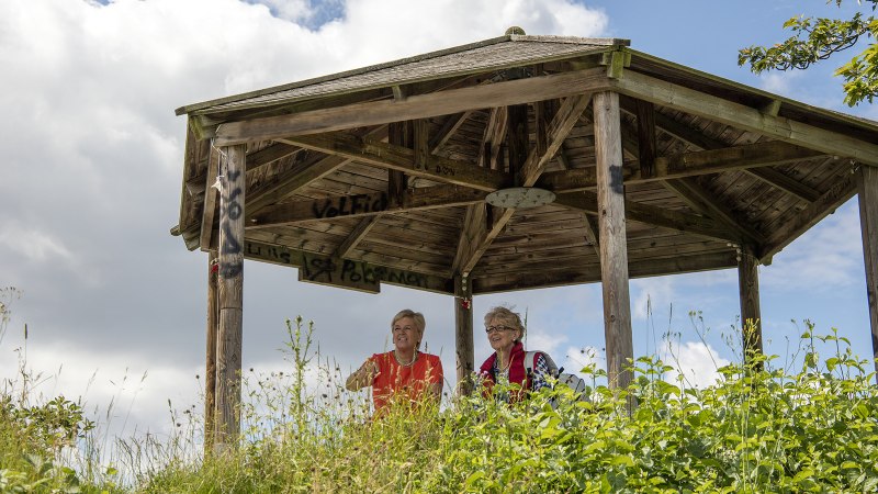 Zwei Personen sitzen in einem hölzernen Pavillon auf einem Hügel, umgeben von Pflanzen und bewölktem Himmel., © SMG, Achim Mende