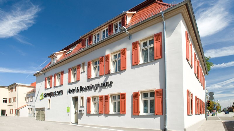Weißes Gebäude mit roten Fensterläden, blauer Himmel. Schriftzug: 'campuszwei Hotel & Boardinghouse'. Moderne Architektur, sonniger Tag., © TOMAS Weißes Gebäude mit roten Fensterläden, blauer Himmel. Schriftzug: 'campuszwei Hotel & Boardinghouse'. Moderne Architektur, sonniger Tag., © TOMAS