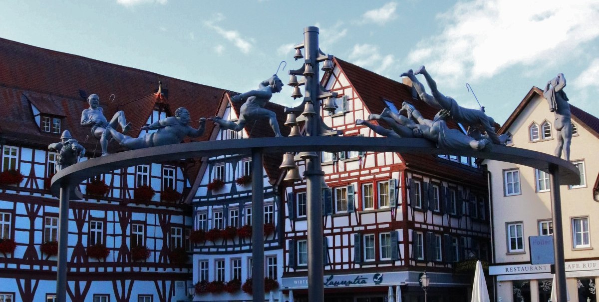 Das Schäferlauf-Denkmal steht auf einem gepflasterten Platz, umgeben von Fachwerkhäusern. Figuren laufen auf einem Metallbogen., © Bad Urach Tourismus