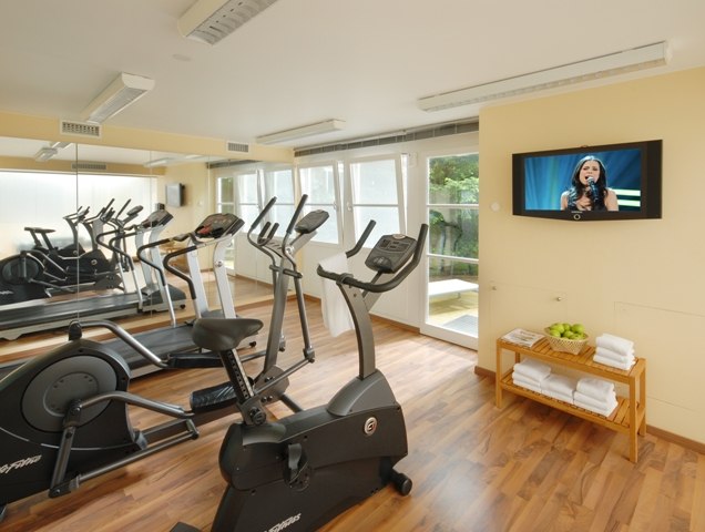 Ein heller Fitnessraum mit Cardiogeräten, Spiegelwand, Fernseher an der Wand und einem Regal mit Handtüchern und Äpfeln., © relexa Waldhotel Schatten Ein heller Fitnessraum mit Cardiogeräten, Spiegelwand, Fernseher an der Wand und einem Regal mit Handtüchern und Äpfeln., © relexa Waldhotel Schatten
