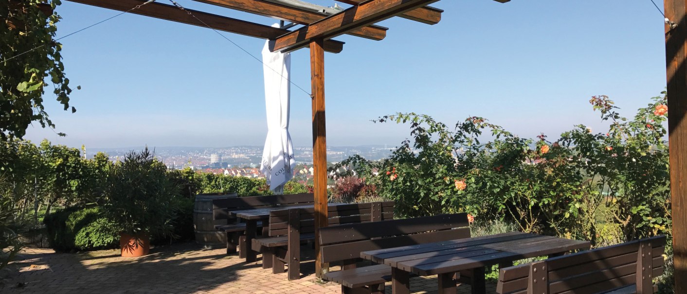 Holzterrasse mit Bänken und Tischen, umgeben von Pflanzen. Im Hintergrund ist eine Stadtlandschaft zu sehen. Sonniges Wetter., © Stuttgart-Marketing GmbH