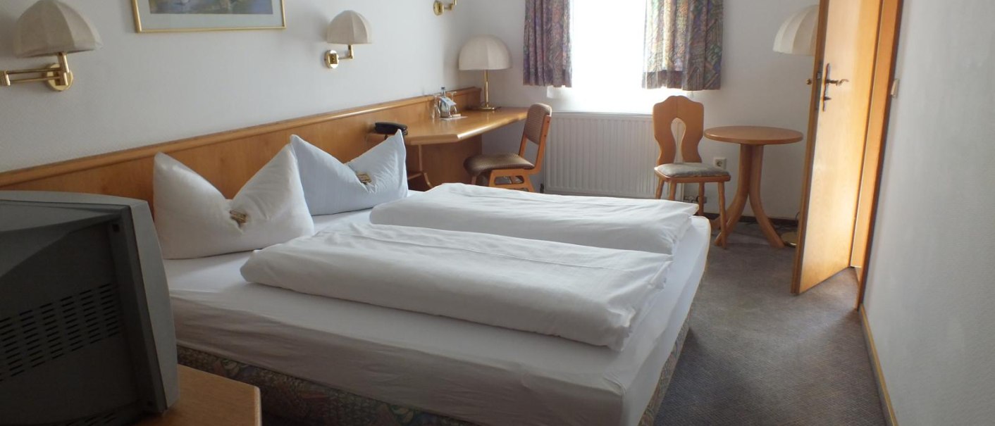 Ein schlichtes Doppelzimmer mit Bett, Schreibtisch, Stuhl und kleinem Tisch. Ein Bild hängt an der Wand, ein Fenster mit Vorhängen lässt Licht herein., © Gasthof zum Bären