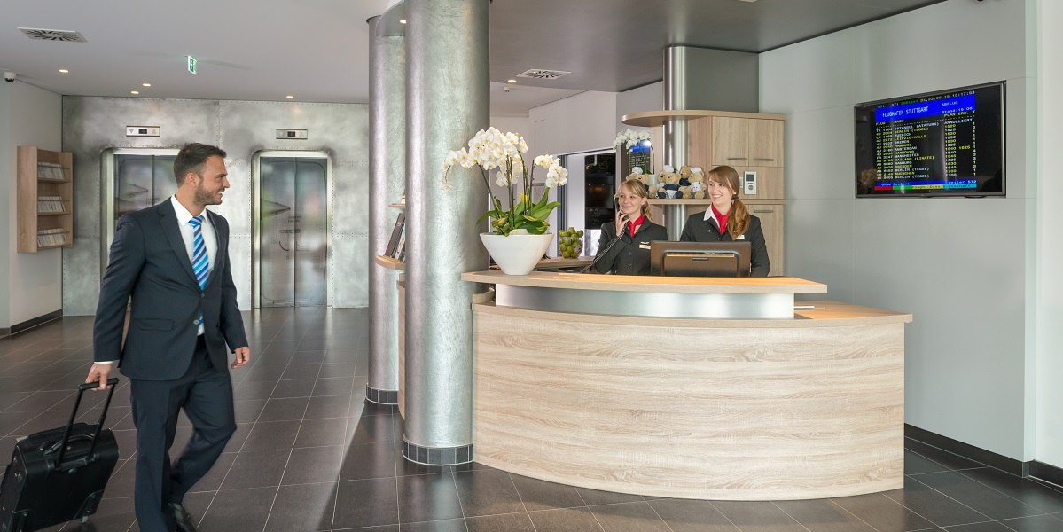 Ein Mann mit Koffer betritt eine moderne Hotel-Lobby. Zwei Rezeptionistinnen stehen hinter einem Empfangstresen. Im Hintergrund sind Aufzüge zu sehen., © Essential by Dorint