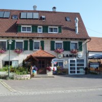 Ein traditionelles Gasthaus mit roten Dachziegeln, grünen Fensterläden und Blumenkästen. Ein Biergarten ist rechts zu sehen.